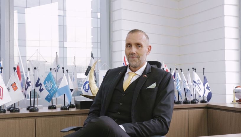 Jo Even Tomren wearing a suit and sitting by his desk. In the background are the flags of different norwegian shipowners. 