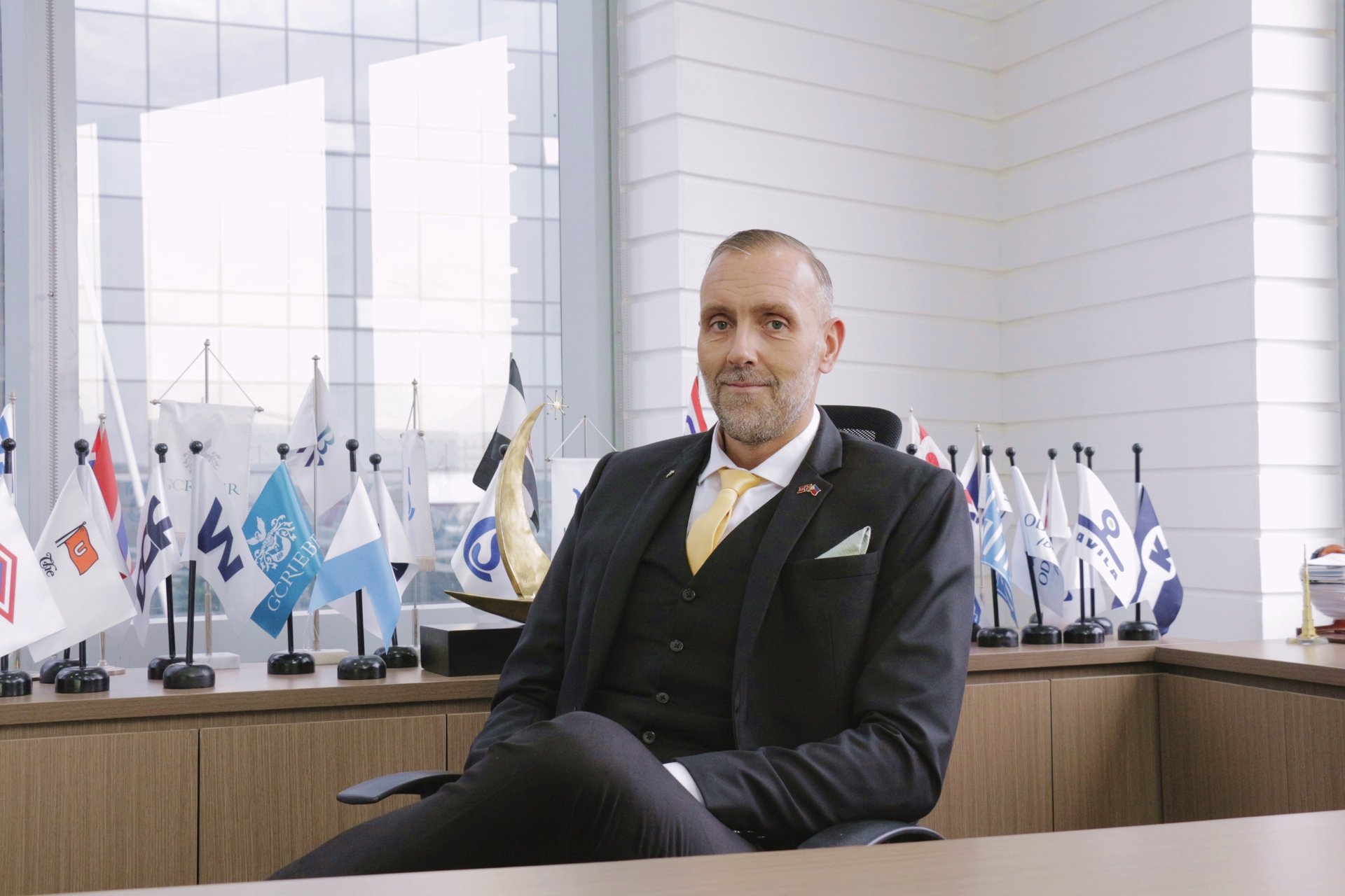 Jo Even Tomren wearing a suit and sitting by his desk. In the background are the flags of different norwegian shipowners. 